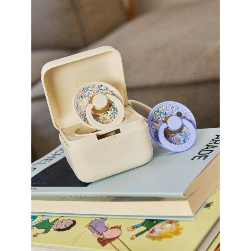 BIBS x LIBERTY Pacifier Box in Chloe Meadow - Ivory Mix and Oscar Meadow - Petal Mix, displayed on stacked books.