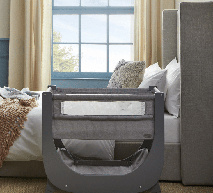 Shnuggle Air Lite Bedside Crib in grey and taupe, positioned next to a bed with soft textiles and natural light.