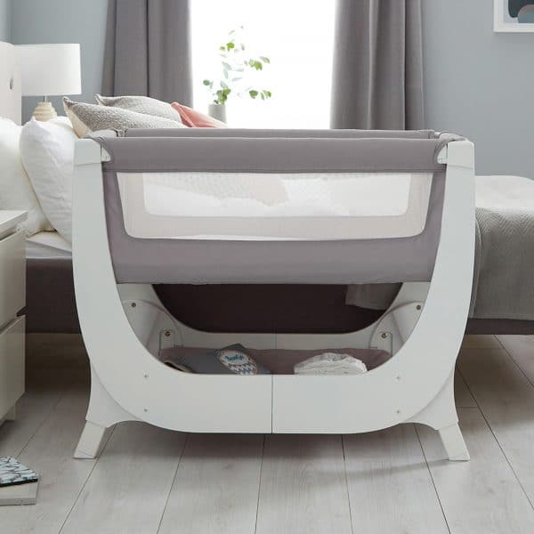 Shnuggle Air Bedside Crib in Dove Grey with a mesh side panel, positioned beside a bed in a cozy nursery setting.