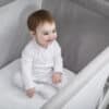 Shnuggle Air Cot Fitted Sheets in use with a smiling baby sitting on the sheet inside a modern cot.
