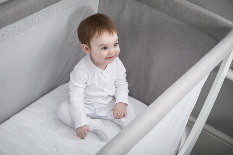 Shnuggle Air Cot Fitted Sheets in use with a smiling baby sitting on the sheet inside a modern cot.