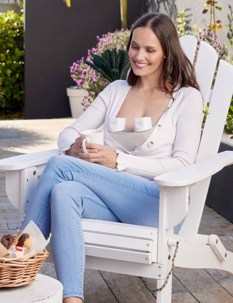 Momcozy Breast Pump S12 Pro in use, worn discreetly under a light cardigan, while a woman relaxes in an outdoor setting.