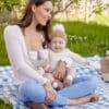 Momcozy Breast Pump S12 Pro held by a smiling mother outdoors, sitting on a picnic blanket with her baby in a cozy outfit.