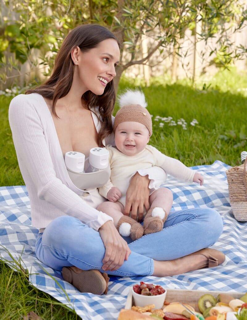 Momcozy Breast Pump S12 Pro held by a smiling mother outdoors, sitting on a picnic blanket with her baby in a cozy outfit.