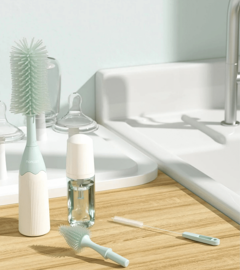 Momcozy Bottle Brush Kit in mint green with a large brush, cleaning solution, and small brush on a wooden countertop.