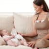 Momcozy Breast Pump S12 Pro worn by a mother sitting on a couch, interacting with her baby in a cozy setting.
