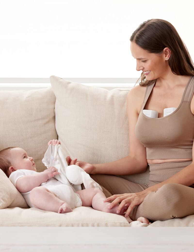 Momcozy Breast Pump S12 Pro worn by a mother sitting on a couch, interacting with her baby in a cozy setting.
