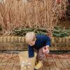 Mushie Silicone Watering Can in Peony held by a child, set in a garden with dried plants and a yellow bucket.