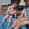 Mushie Silicone Watering Can in Peony, held by a child sitting by a pool, with another child nearby.