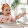 Back to Nature Wood Ring Rattle in blue and pink, held by a smiling baby in a high chair, promoting sensory play.