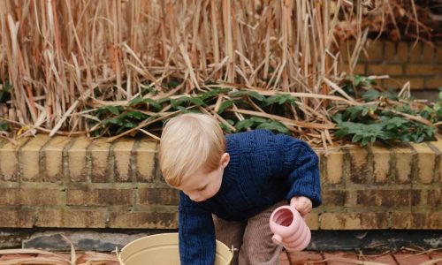 Mushie Silicone Watering Can in Peony held by a child, set in a garden with dried plants and a yellow bucket.